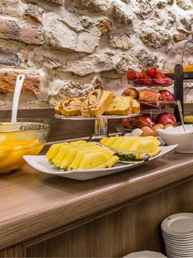 Porcjowane plastry ananasa, ciasto drożdżowe, miska z pokrojonym mango i kolekcja jabłek i truskawek na drewnianym blacie z kamienną ścianą w tle.