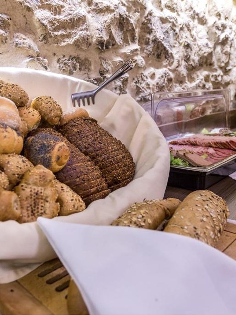 Kosz z różnymi rodzajami pieczywa, w tym bułki i chleby, oraz tacka z plasterkami wędliny na tle kamiennej ściany.