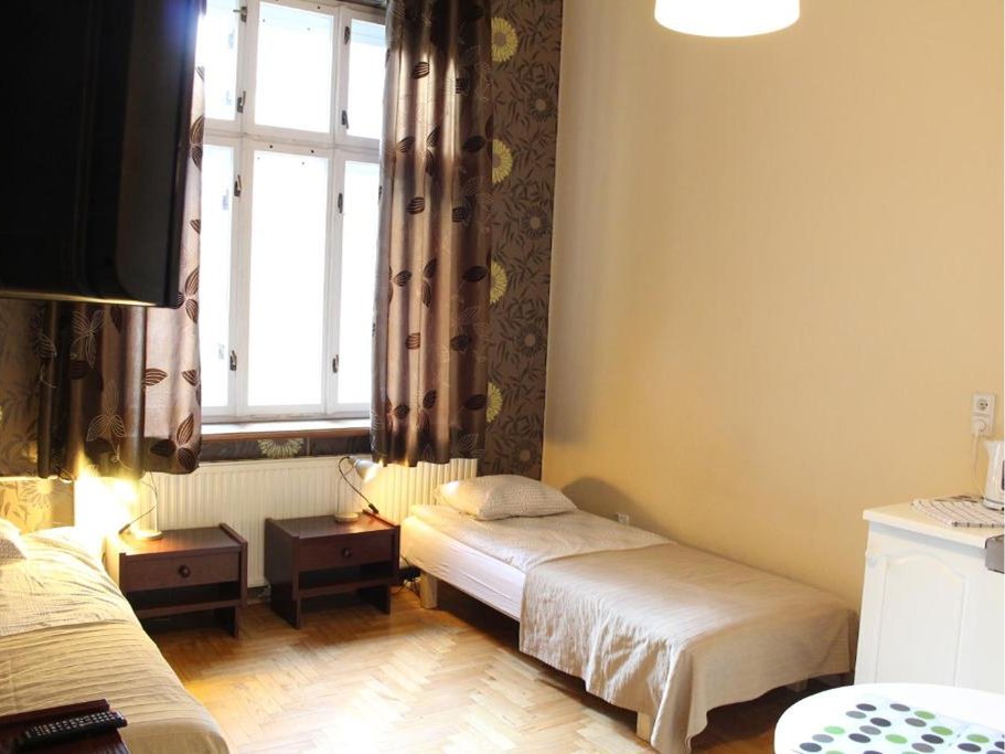 Pokój z pojedynczym łóżkiem i sofą, dwoma nocnymi stolikami, oknem z zasłonami oraz jasnymi ścianami i podłogą drewnianą.