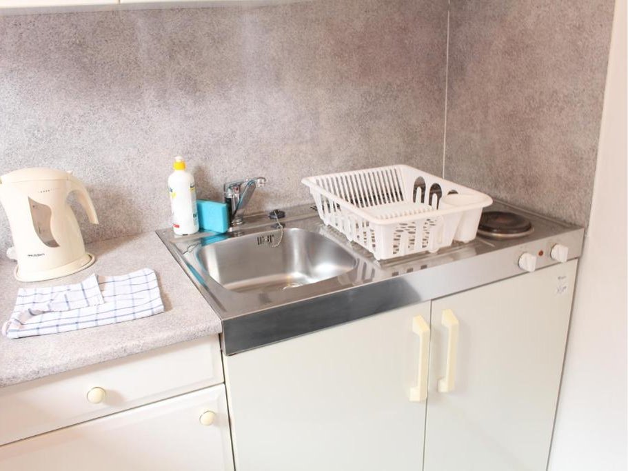 Mała kuchnia z metalowym zlewem, suszarką na naczynia, czajnikiem elektrycznym i dwupalnikową kuchenką elektryczną.
